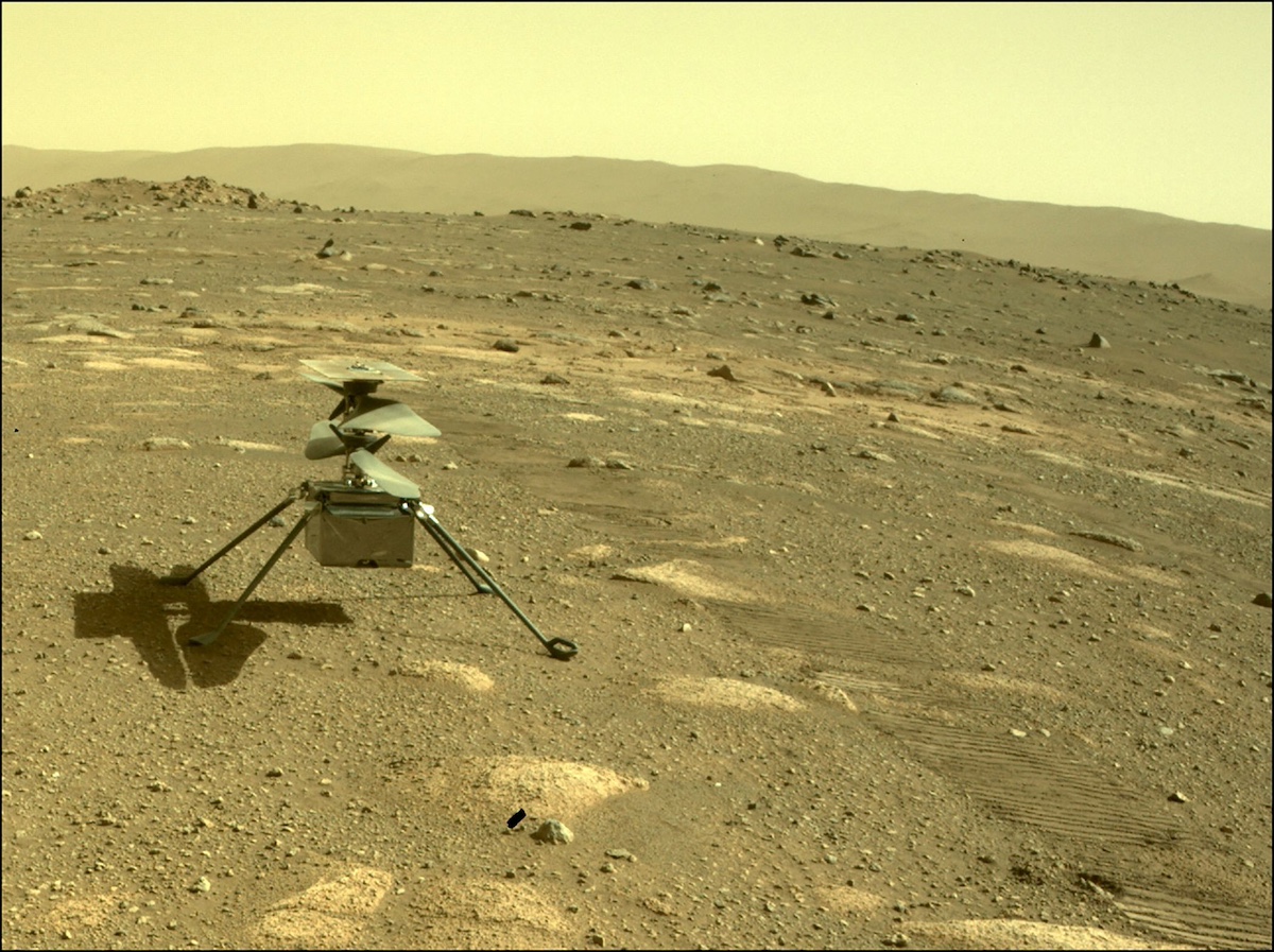 The image shows a small helicopter-like drone on a rocky, dusty landscape under a yellow-brown sky, resembling the surface of Mars.