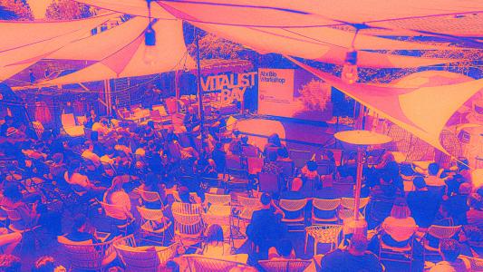 A large group of people sits outdoors under shade sails at Vitalist Bay, facing a stage with a sign that reads "Vitalist Bay" and "Alt Bio Workshop.