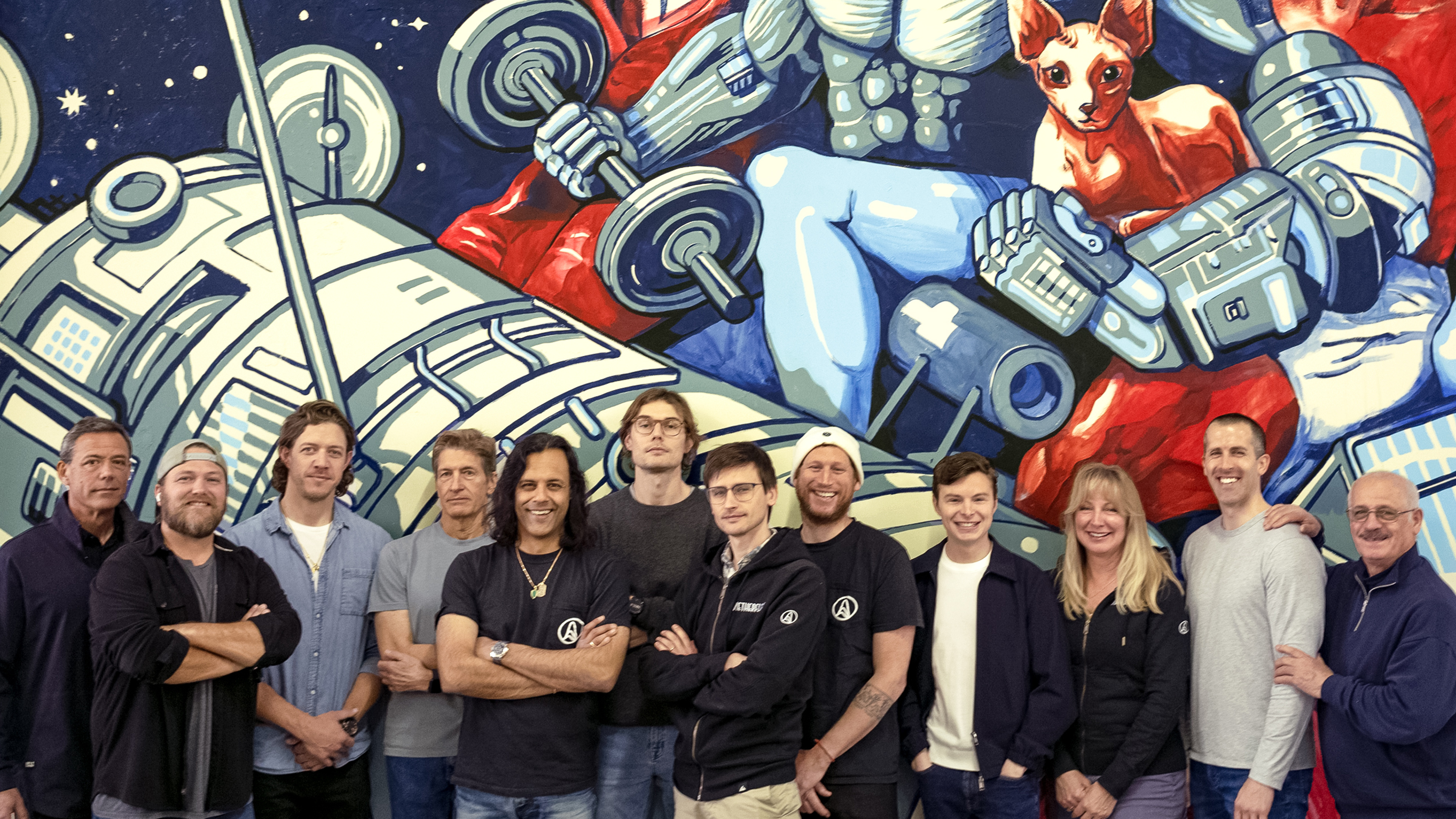 A group of thirteen people stand in front of a colorful mural featuring a futuristic robotic scene with a red cat and mechanical elements.