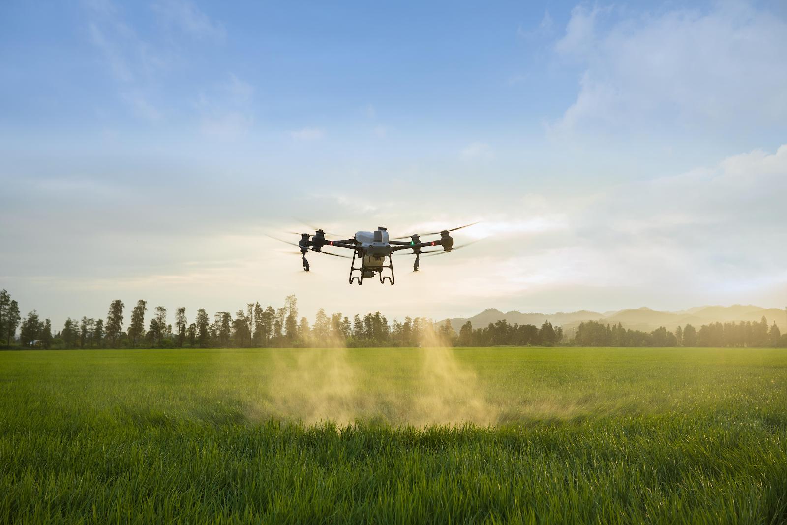 A drone hovers over a green field, dispersing a mist, possibly for agricultural purposes, with a background of trees and distant mountains during daytime.