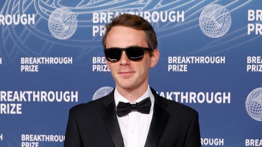 A man in a black tuxedo and sunglasses stands in front of a blue backdrop with the Breakthrough Prize logo and text.