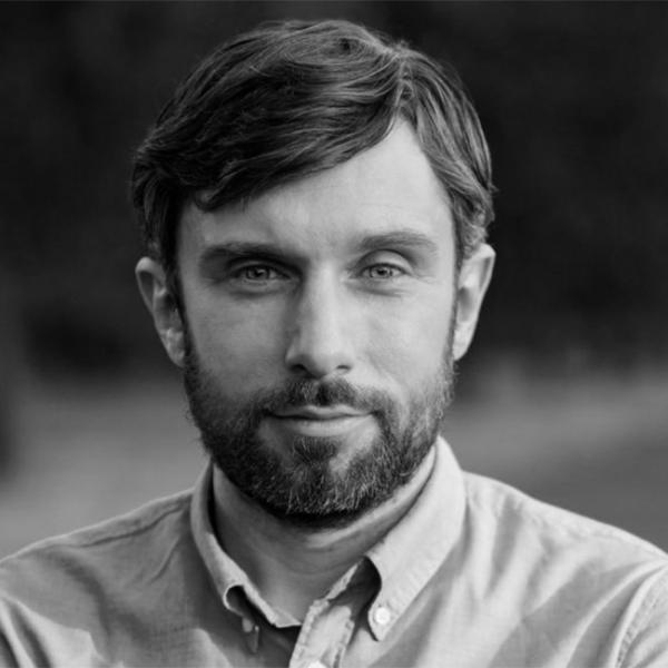 A man with short brown hair and a beard, wearing a button-up shirt, looks directly at the camera in an outdoor setting. The image is in black and white.