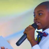 A young person in a school uniform speaks into a microphone while holding a piece of paper.