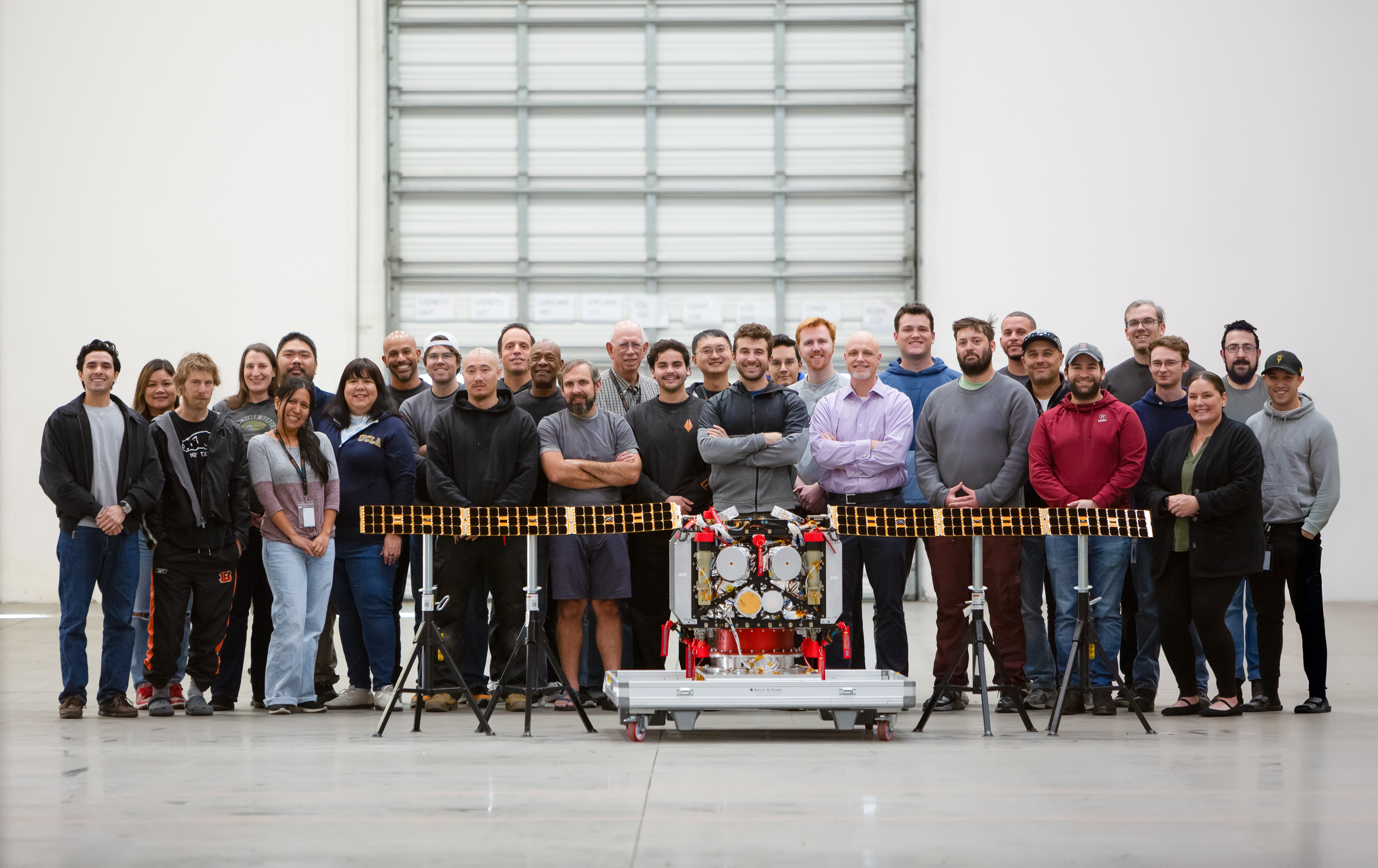 A group of people poses indoors behind a robotic device with extended solar panels, positioned in front of a large, closed industrial door.