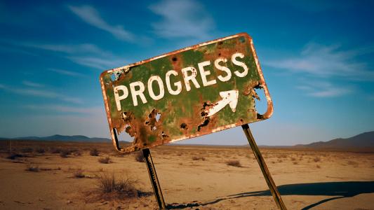A heavily rusted and damaged sign reading "PROGRESS" with an arrow stands tilted in a barren desert landscape under blue sky.