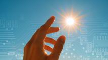 A hand reaches toward the sun against a blue sky, with faint digital circuit patterns overlaid in the background.