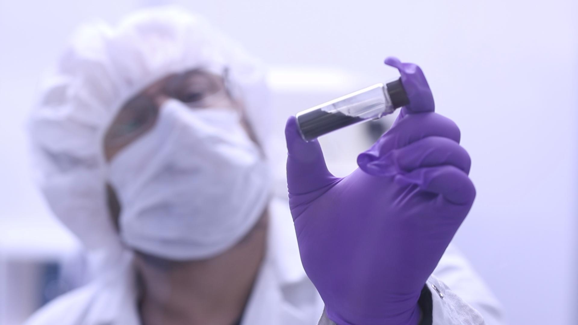 A scientist wearing protective clothing and purple gloves holds up a test tube for examination in a laboratory setting.