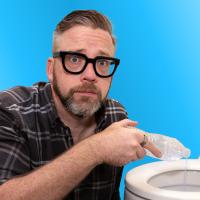 A man with glasses pours liquid from a plastic bag into a toilet against a blue background.