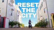 A person rides a bicycle between two modern white buildings under a clear blue sky, with large text reading "RESET THE CITY" above.