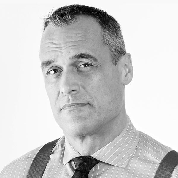 A middle-aged man with short gray hair wearing a striped dress shirt, tie, and suspenders, looking directly at the camera against a plain background.