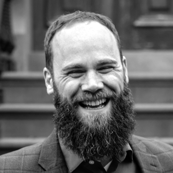 A bearded man wearing a plaid jacket smiles broadly, showing his teeth. The background is blurred steps and a wooden door. The image is in black and white.