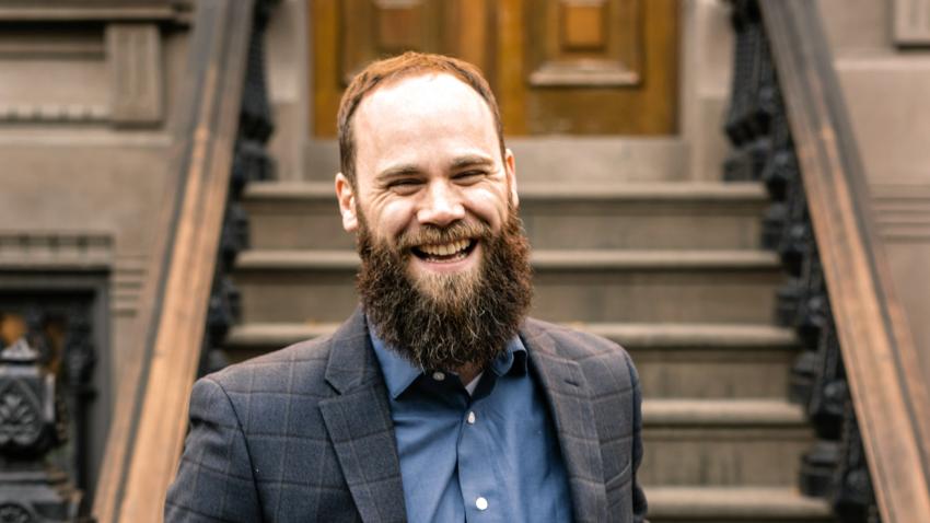 Daniel Jeffries in a plaid suit jacket and blue shirt smiles while standing in front of a brownstone staircase.