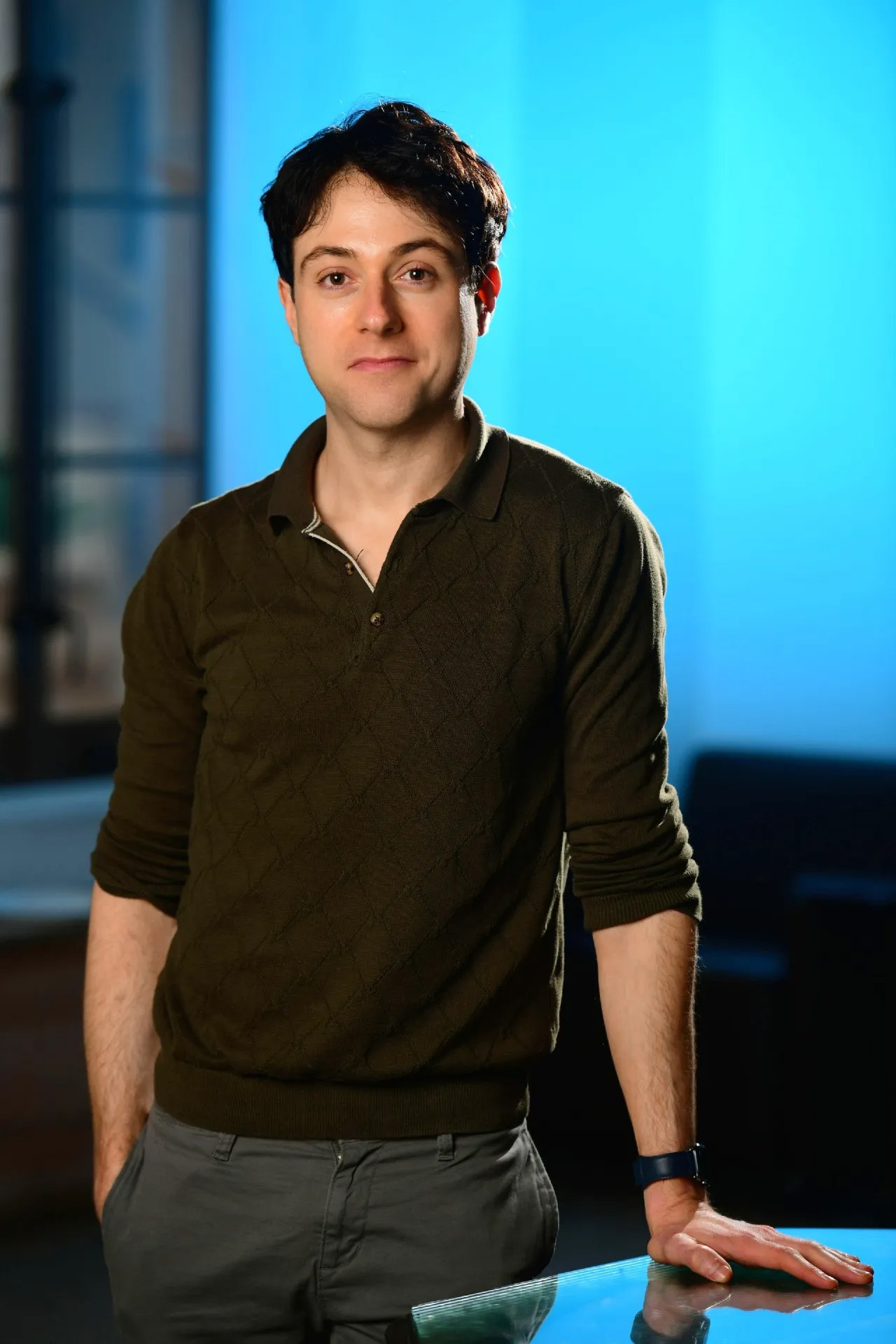A person with short dark hair wearing a dark green sweater and gray pants stands indoors, resting one hand on a glass surface—perhaps contemplating ways to cure coma. The background is softly lit with blue tones.