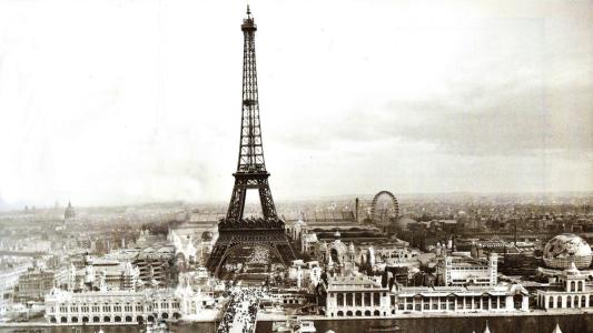 Black and white photo of the Eiffel Tower during the 1889 Exposition Universelle in Paris, with crowds, pavilions, and a Ferris wheel in the background.