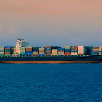 A large cargo ship is sailing on the ocean, loaded with colorful shipping containers against a backdrop of a clear sky and distant land.