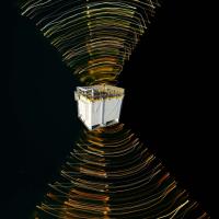 A cube-shaped satellite is surrounded by swirling lines of light in space against a black background, illustrating electromagnetic fields or signal patterns.