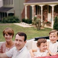 A family of four is seated in a red convertible car, parked in front of suburban houses. The parents sit in the front, while the two children sit in the back.