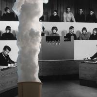 A black and white TV game show set with an audience and four contestants at desks. A cloud of smoke or steam rises from an object in the foreground.