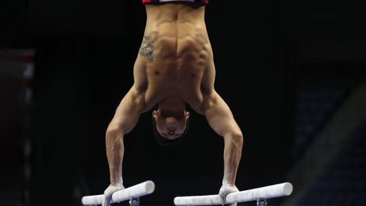 A male gymnast performs a handstand on parallel bars in a competition setting, displaying upper body strength and control.