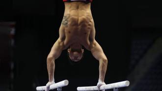 A male gymnast performs a handstand on parallel bars in a competition setting, displaying upper body strength and control.
