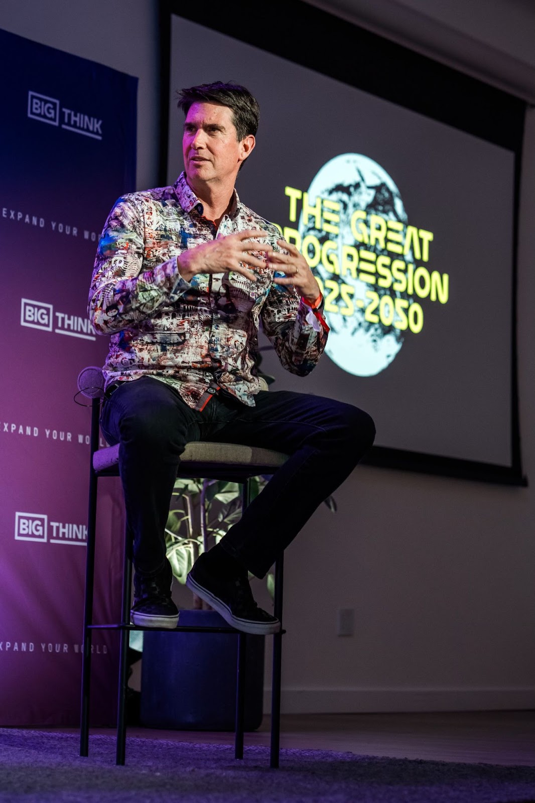 A man sits on a stool speaking, with a screen behind him displaying the text "THE GREAT PROGRESSION 2025-2050" and the Big Think logo visible on banners.