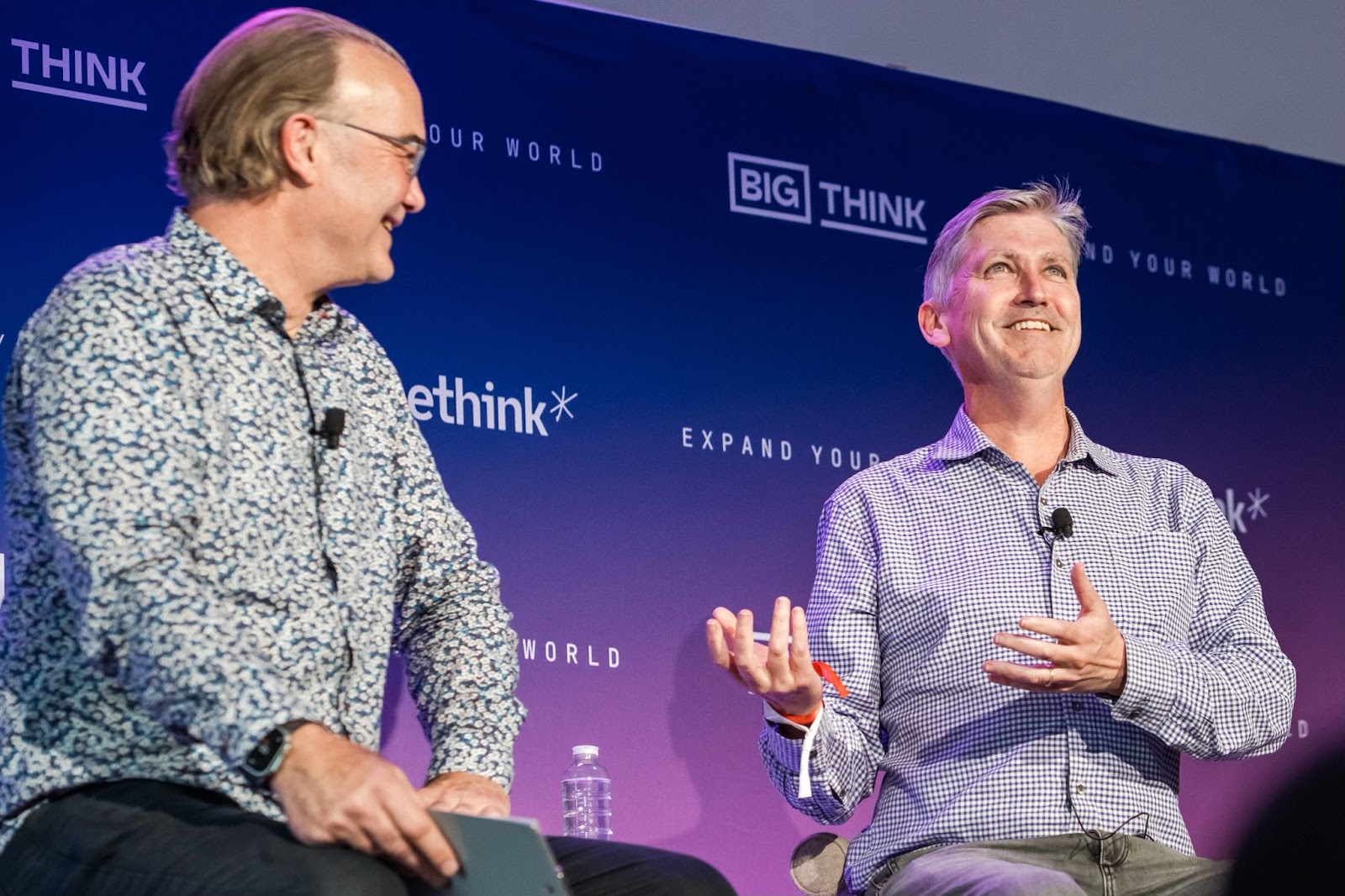 Two men sit and talk on stage at a Big Think event, one holding a notebook and the other gesturing with his hand.