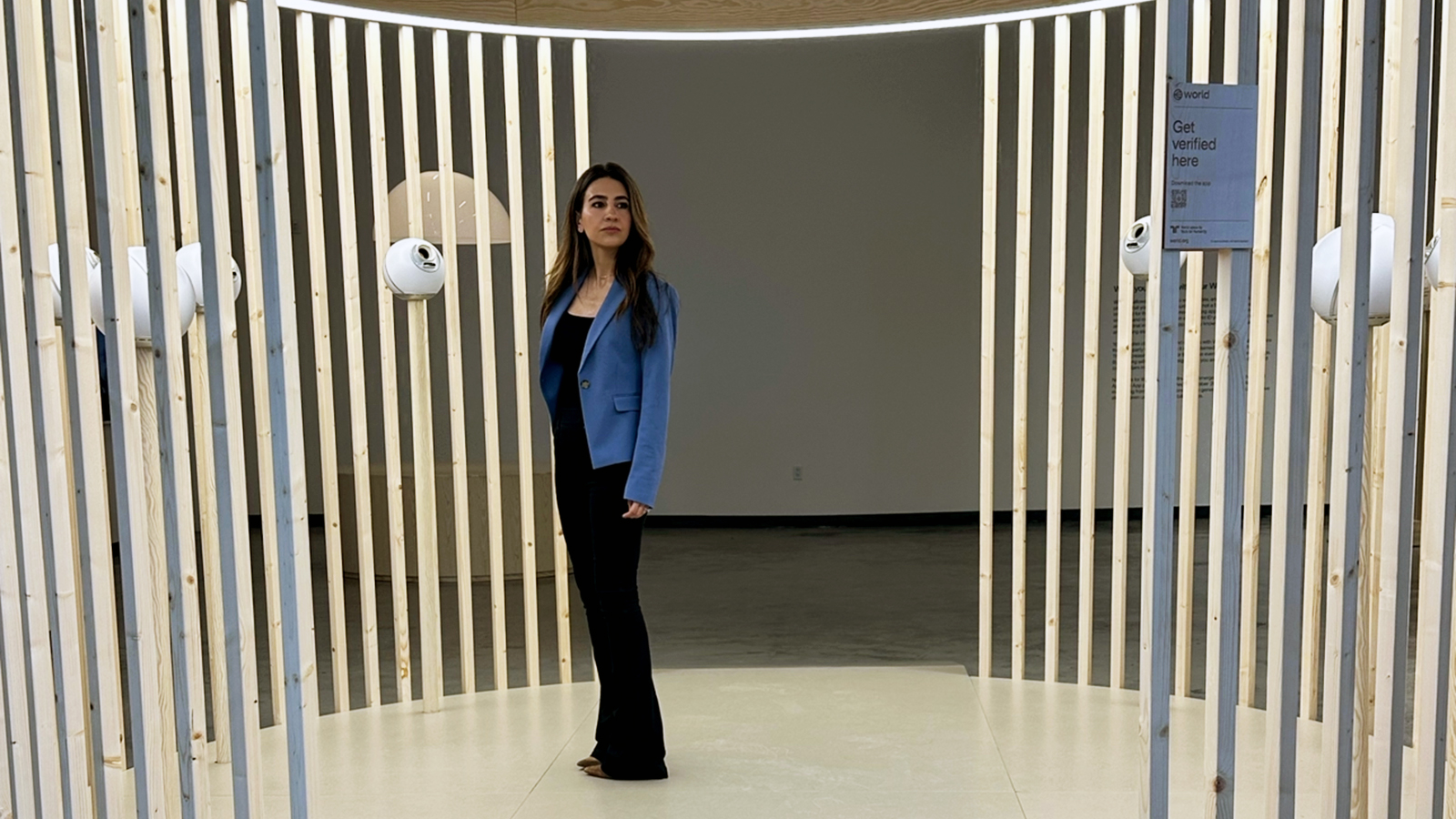 A woman in a blue blazer stands inside a circular structure made of vertical wooden slats, with lights overhead and a sign on the right reading "Get verified here.