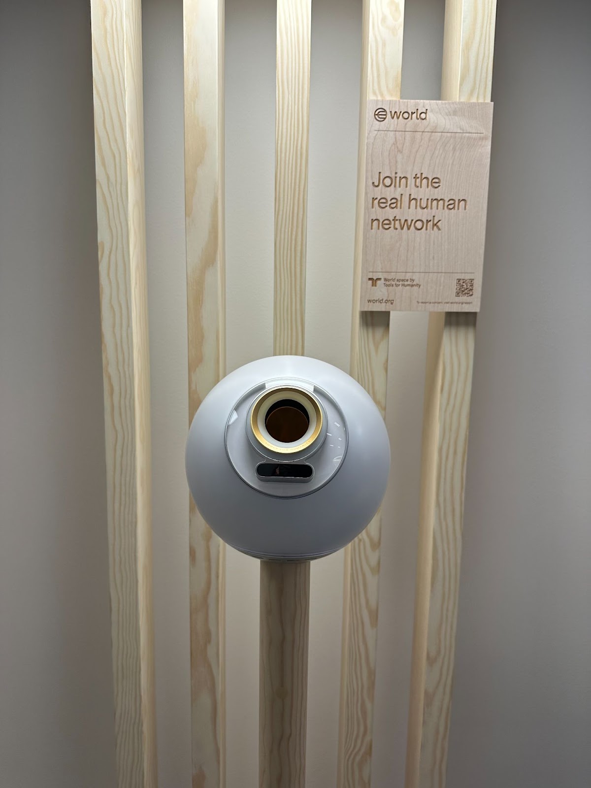 A white spherical device mounted on vertical wooden slats, with a sign above reading "Join the real human network" and a QR code on the right side of the sign.