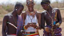 Three people in traditional attire look at a smartphone together outdoors, holding long sticks decorated with colorful patterns.