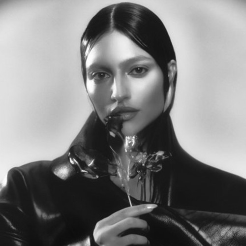 A woman with straight dark hair and a serious expression holds a flower, wearing a dark, shiny coat against a soft, light background.