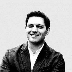 Black and white photo of a man with short hair wearing a blazer and shirt, smiling and looking slightly to the right.