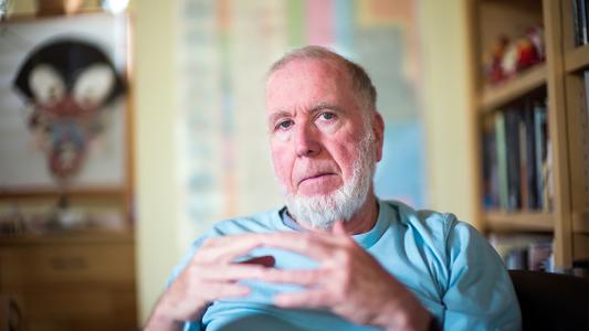A man with a gray beard and wearing a light blue shirt sits with his hands clasped, in a room with shelves and artwork in the background.