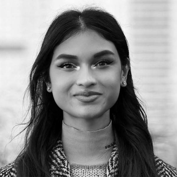 A young woman with long dark hair and earrings looks at the camera, wearing a high-neck sweater and patterned coat. The image is in black and white.
