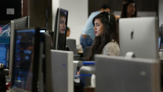 A woman with long dark hair works at a computer in an office, surrounded by multiple monitors and other people in the background.