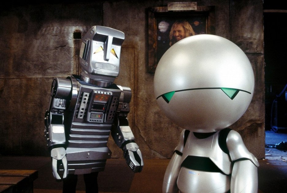 Two robots stand in a dimly lit room with a framed portrait on the wall behind them; one robot is boxy and silver, the other has a round head and green eyes.