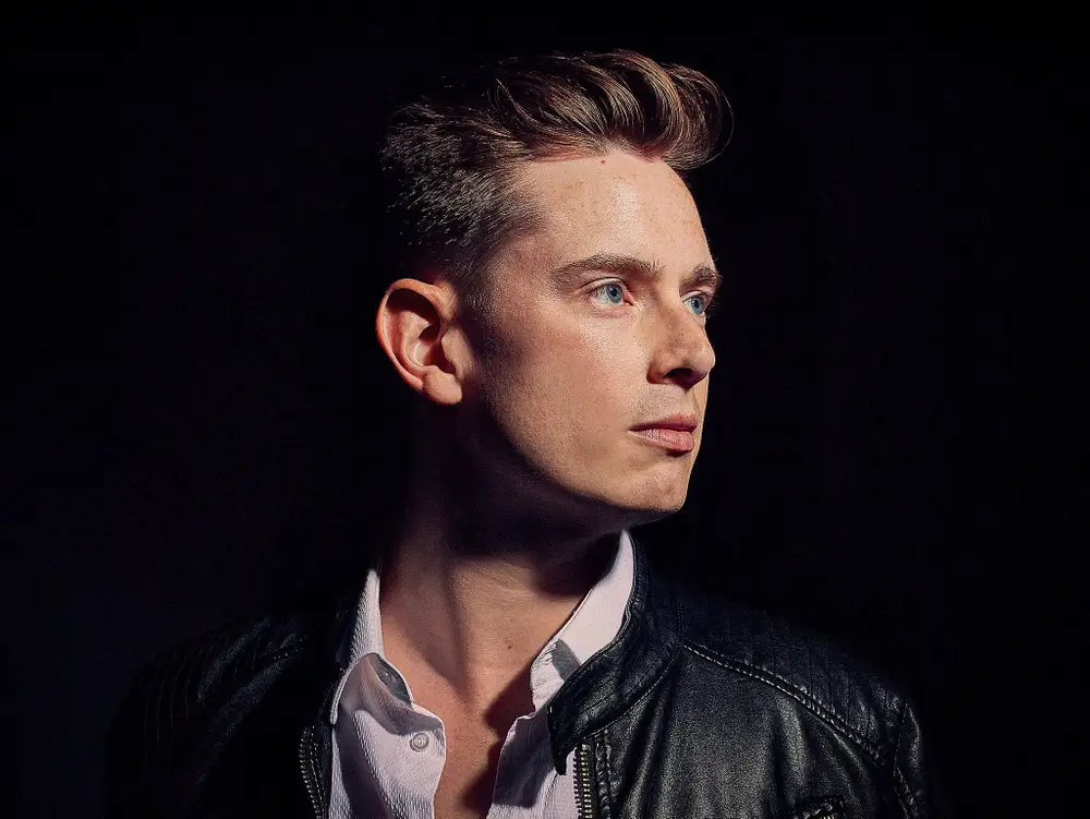 A person with short hair looks to the side, wearing a leather jacket and a white shirt, set against a dark background.