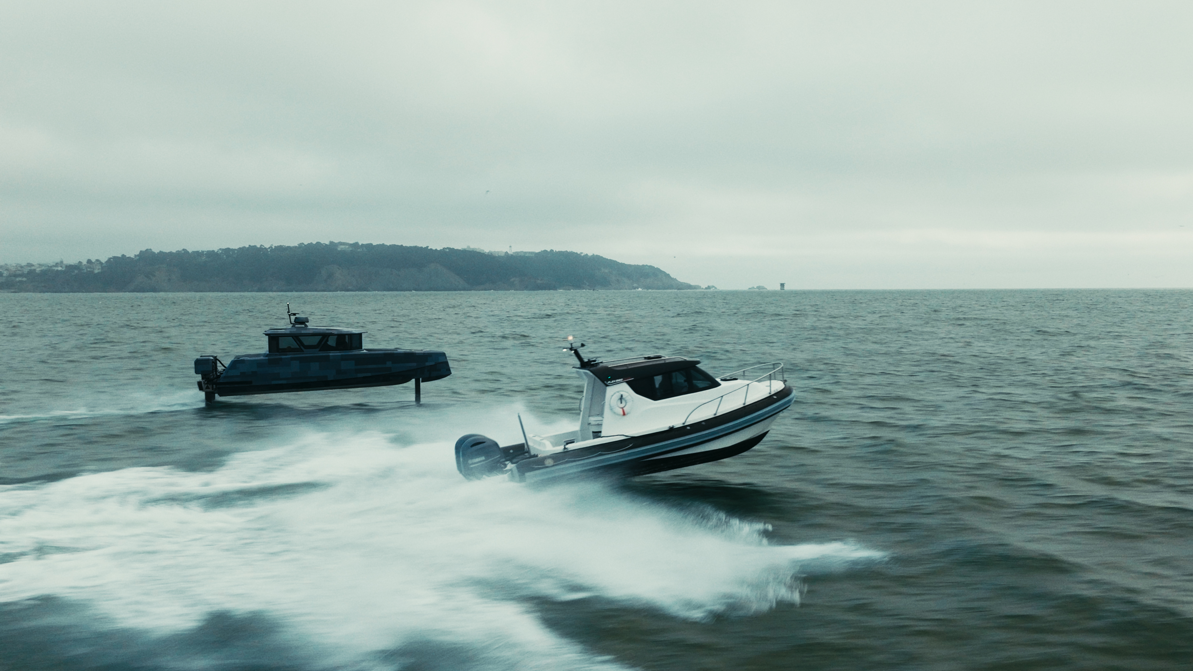 Two motorboats speed across open water under an overcast sky, with a coastline and hilly land visible in the background.