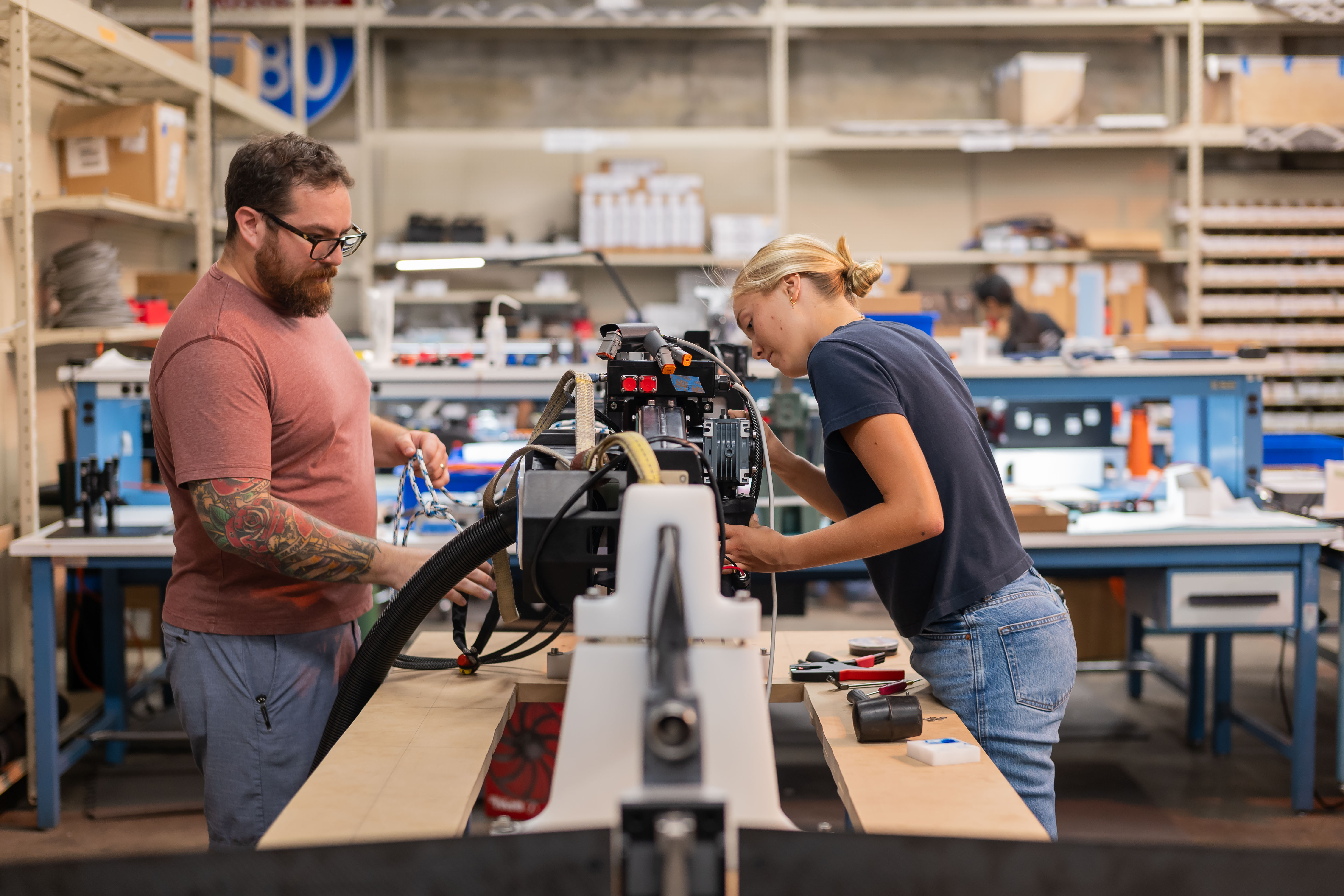 Two people work together on a machine in an industrial workshop, surrounded by tools and equipment on shelves and workbenches.