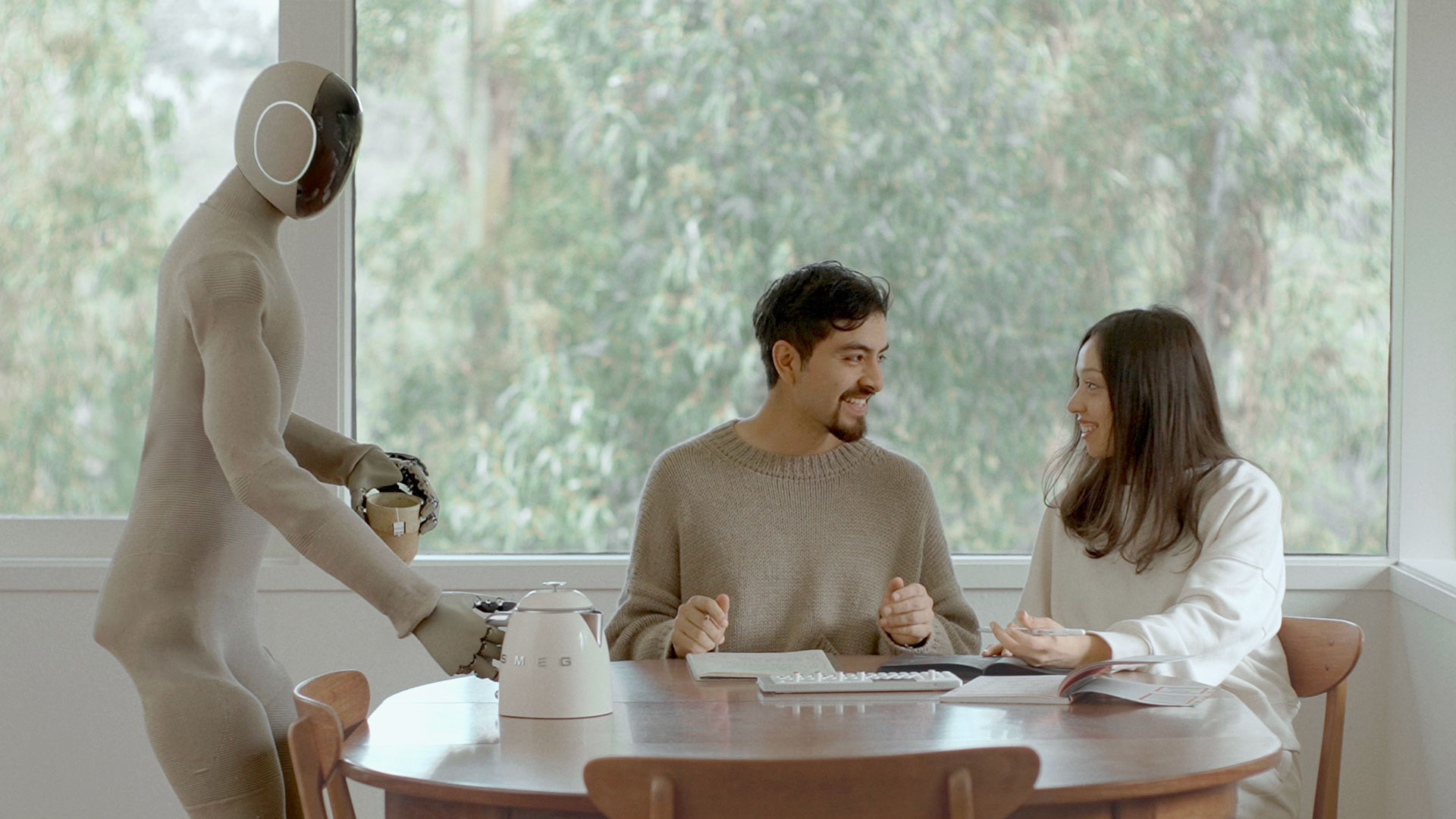 A humanoid robot serves coffee to a man and woman sitting at a round wooden table by a large window.