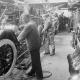 Workers assemble cars in an early 20th-century automotive factory, surrounded by vehicle parts and tools.