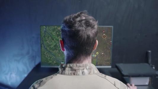 A person in military uniform sits at a desk, looking at a computer monitor displaying a digital map with various symbols and markings.