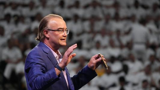 Peter Leyden, wearing a blue suit and glasses, speaks and gestures on a stage about the Great Progression to a large audience clad in white.
