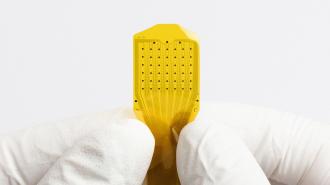 A gloved hand holds a small, yellow electronic microchip with a grid of black dots on its surface against a plain white background.
