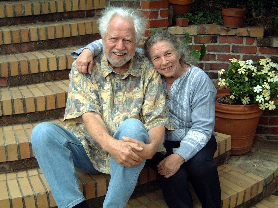 Older couple sitting on brick steps with potted flowers, the man wearing a patterned shirt and jeans, the woman in a blue blouse and black pants, both smiling at the camera.