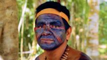 Individual with blue and red face paint, wearing a yellow headband and a necklace, in an outdoor wooded area.