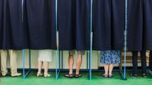 Six individuals standing behind closed blue curtains in voting booths, with only their legs and feet visible, suggesting they are engaged in the voting process.