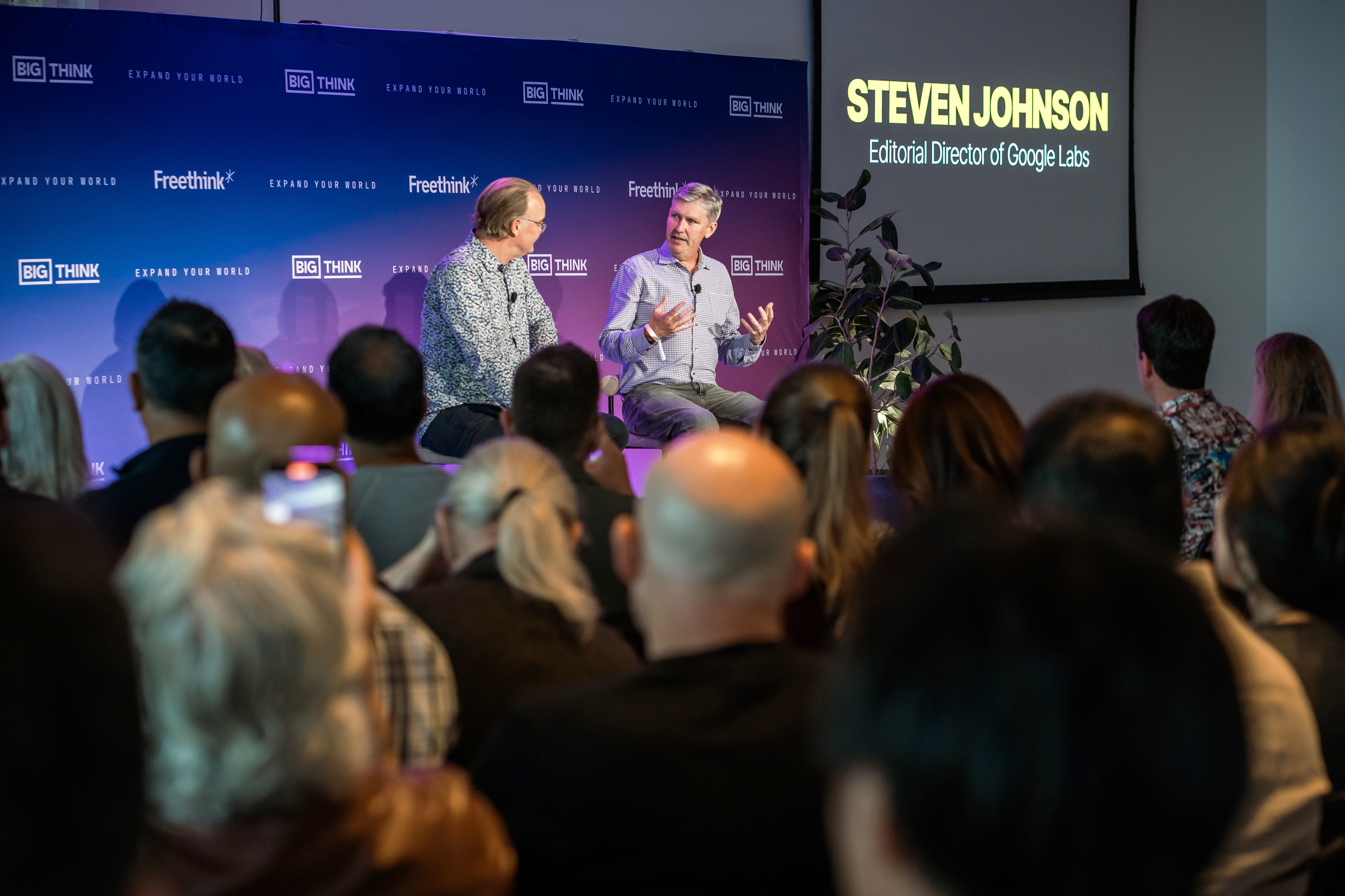 Two men sit on stage having a discussion in front of an audience; a screen displays "Steven Johnson, Editorial Director of Google Labs.