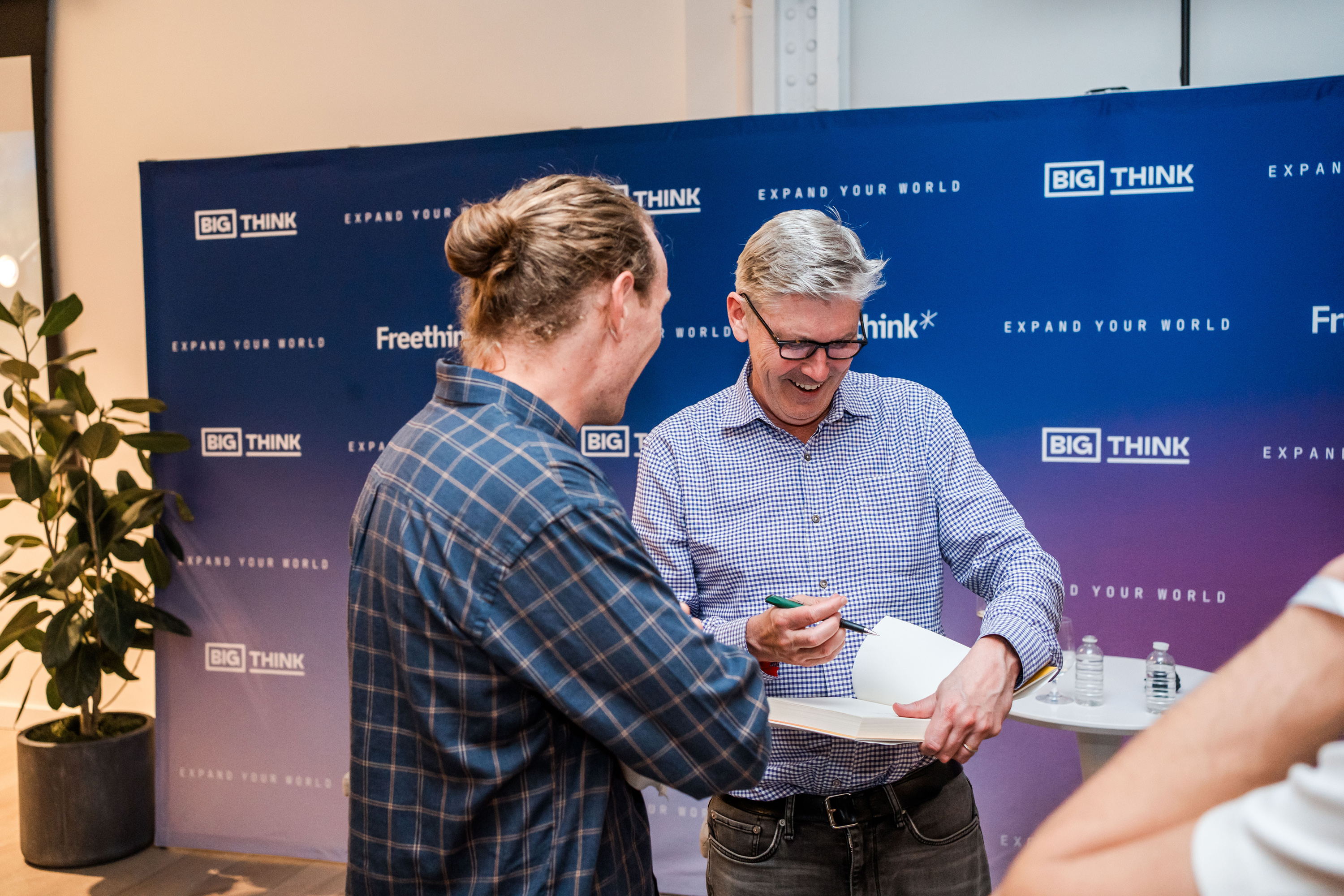 Two men stand at a table as one signs a paper; a "Big Think" event backdrop is in the background.