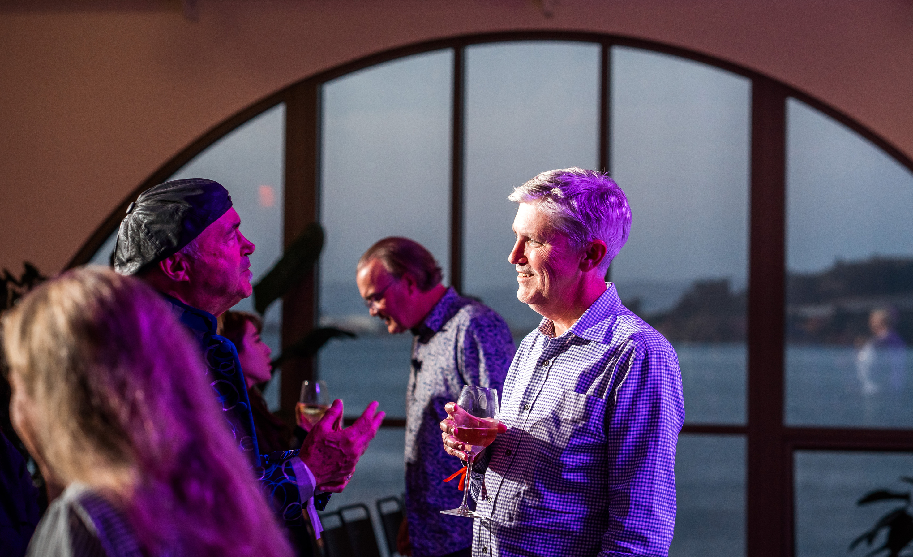 A group of people stand and talk near large arched windows overlooking water; one man in front holds a drink and smiles.