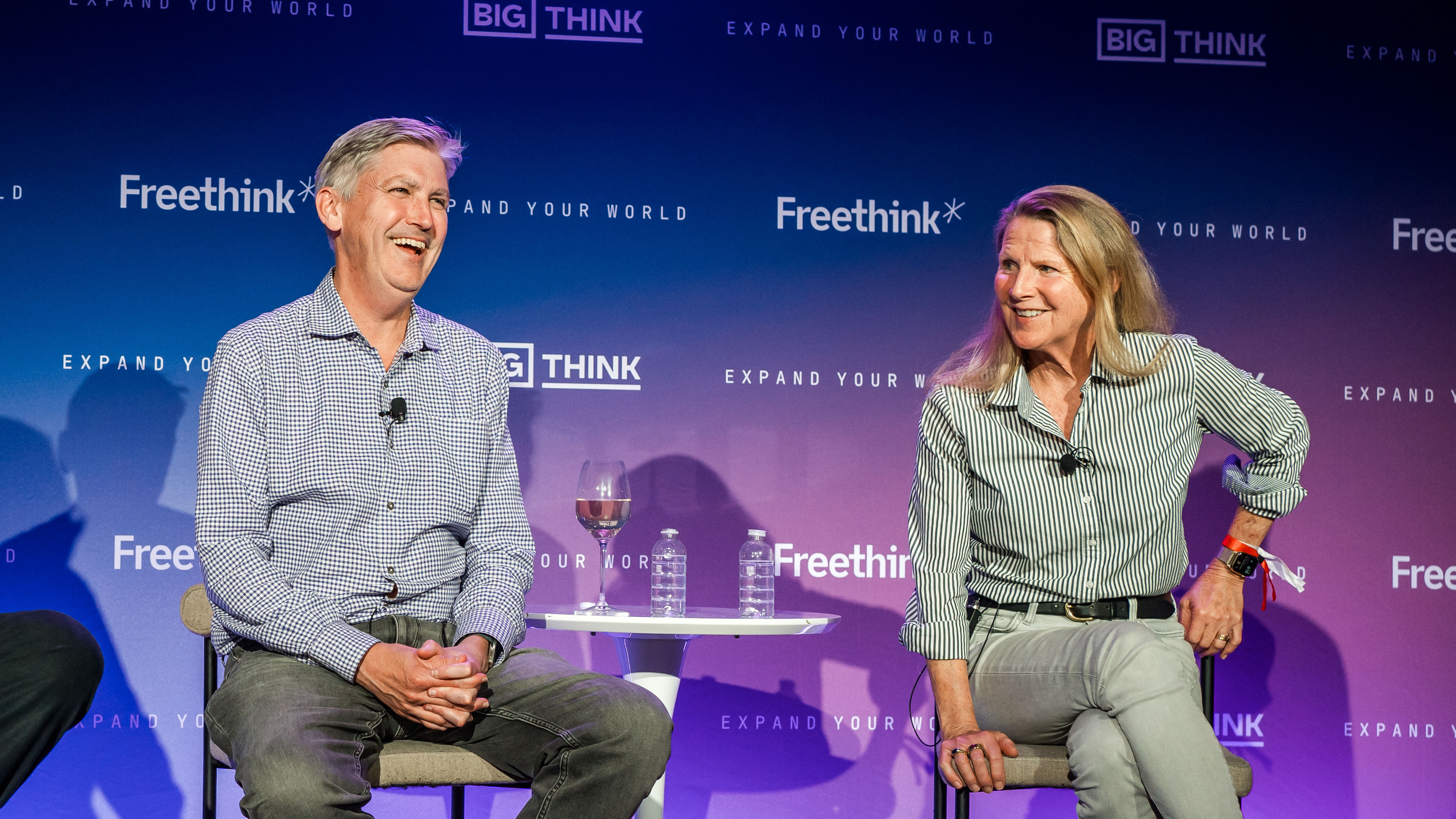 Two people sit and talk on stage at a Freethink and Big Think event, with water glasses and a wine glass on a small table between them.